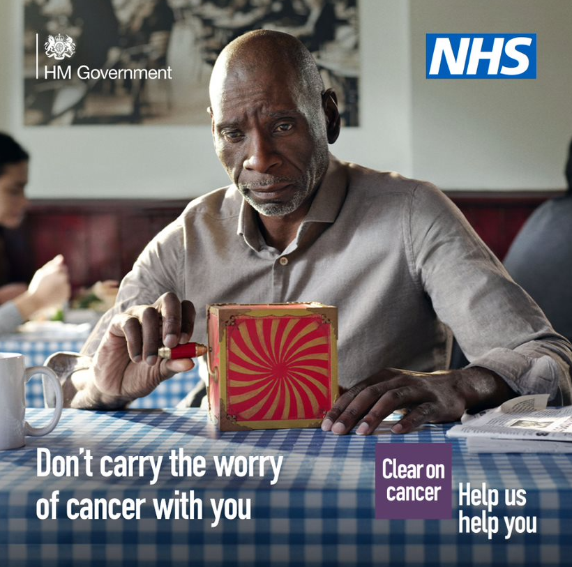 A middle-aged person sitting a cafe with a red jack-in-the-box toy on the table across from them. The person is looking at the red-jack-in-the-box with a concerned expression on their face. The text reads: Don’t carry the worry of cancer with you. The NHS logo is in the top right hand corner.
